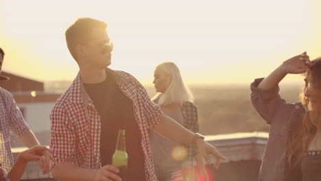 A-young-russian-man-in-trendy-glasses-with-beer-moves-in-a-dance-on-the-party-with-his-friends-on-the-roof.-He-moves-in-dance-with-two-girls.
